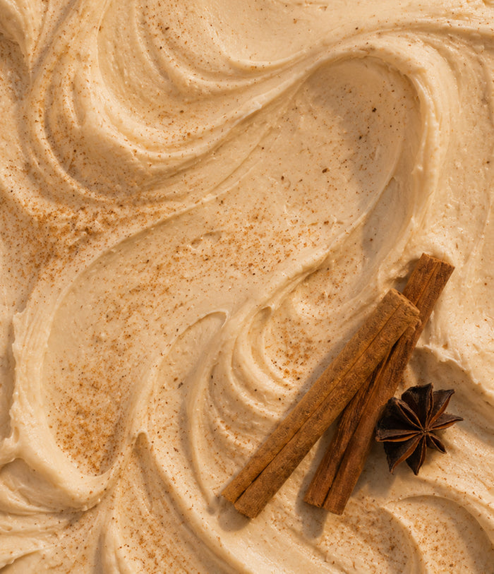 Creamy beige whipped cream with swirls and a light brown spice dusting, styled with two cinnamon sticks and a star anise on a close-up background.
