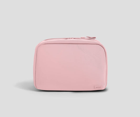 A light pink zippered toiletry bag with rounded corners and a small rectangular "Away" brand tag on the bottom right, shown against a solid light gray background.