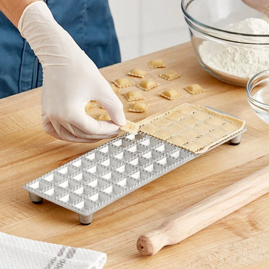 A person using the Imperia RavioChef ravioli mold to shape pasta dough into ravioli, with a wooden rolling pin and flour in the background.